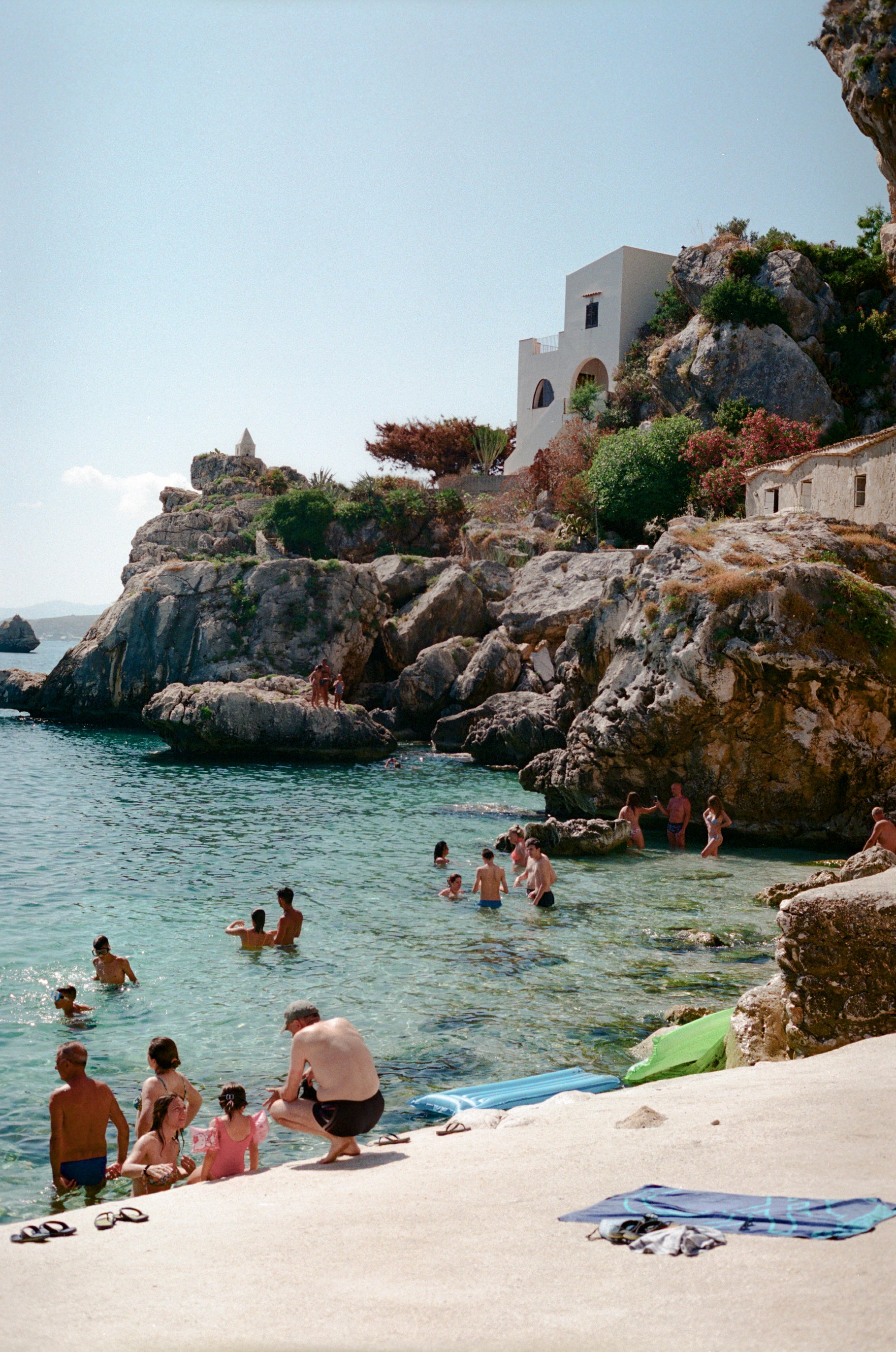 Photograph of swimmers in the sea in Scopello, Sicily, Italy. WIth a white washed house in the cliffside.