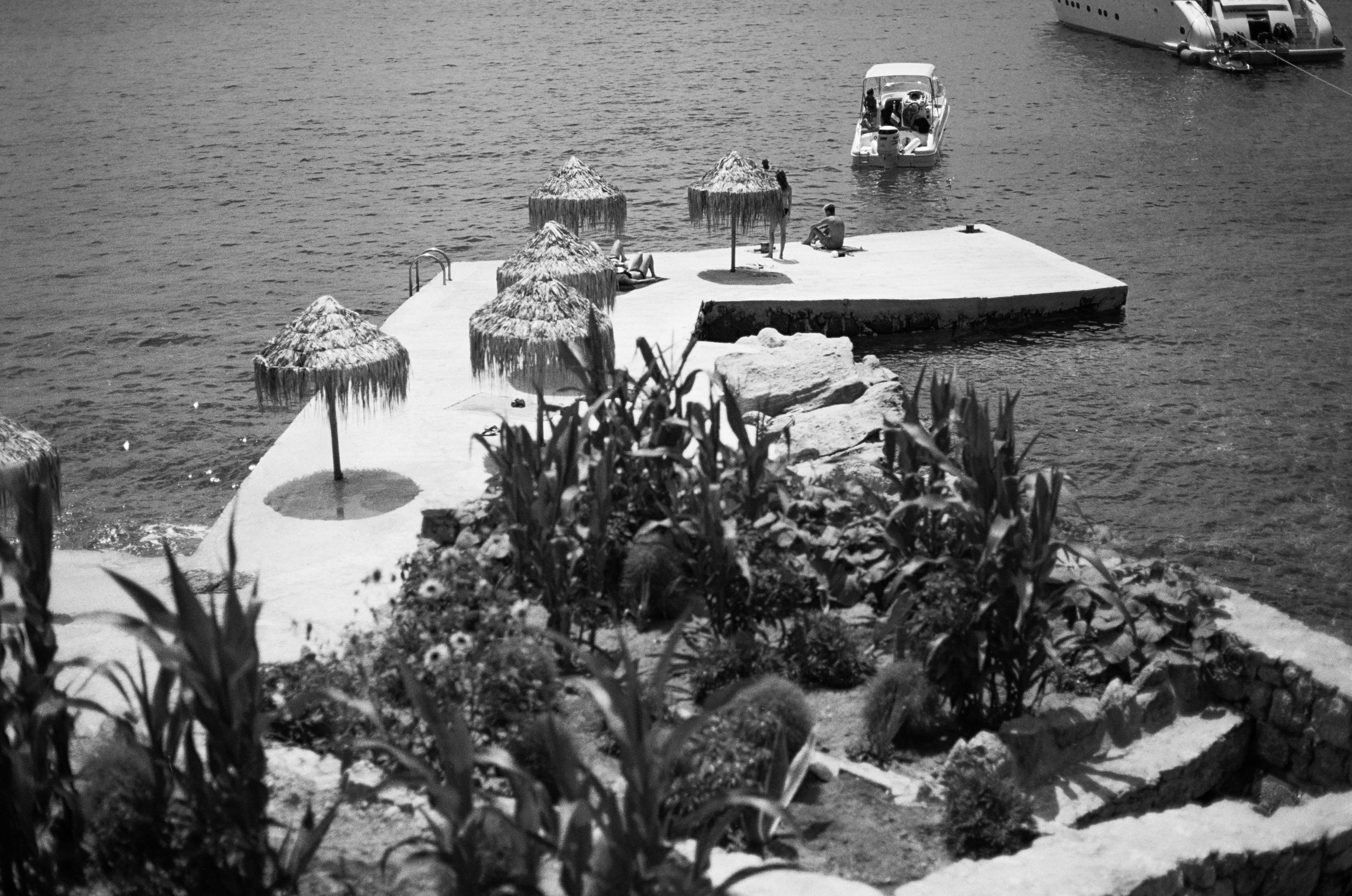 Photograph of beach goers laying on a pier with umbrellas by the sea in Mykonos. With a boat docked in the background and native plants in the foreground.
