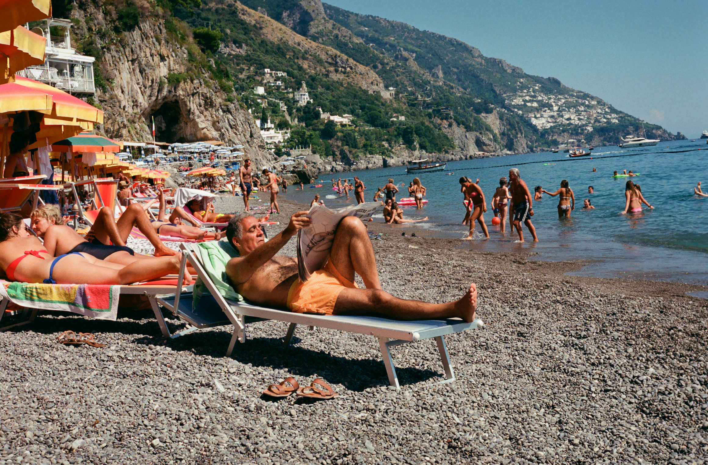 Photograph of an elderly man reading the newspaper on a sun lounge on the iconic Positano shoreline.