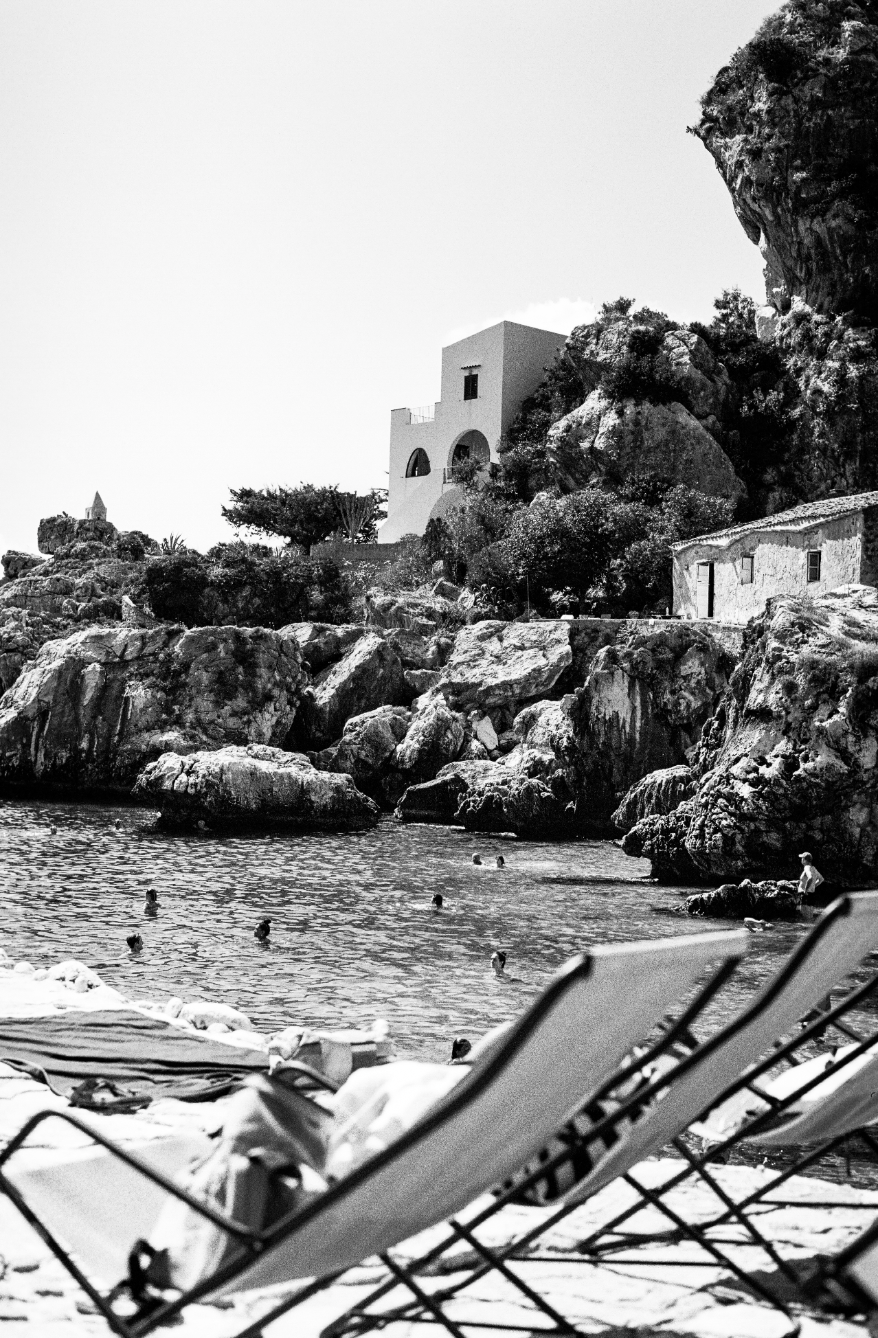 Photograph of Tonarra di Scopello, an old tuna fishing port now a local swimming spot. White wash buildings in the cliffside in the background and sun lounges in the foreground.