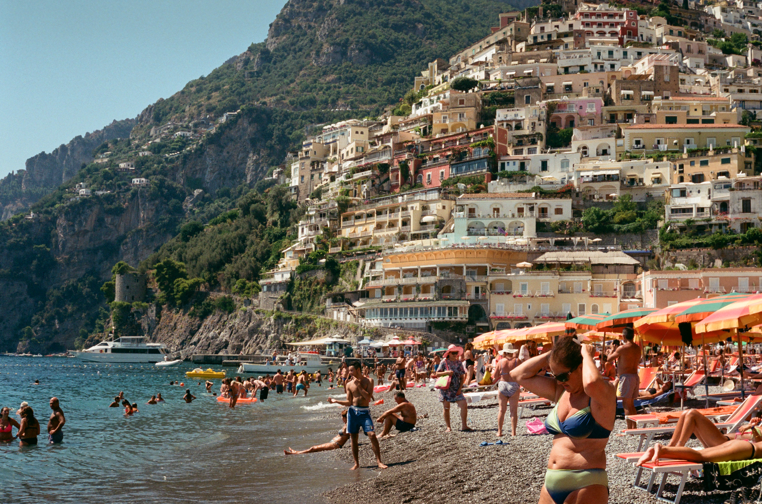 Photograph of the iconic Positano main beach. The famous landacape of cliffside buildings with sea and beach-goers.