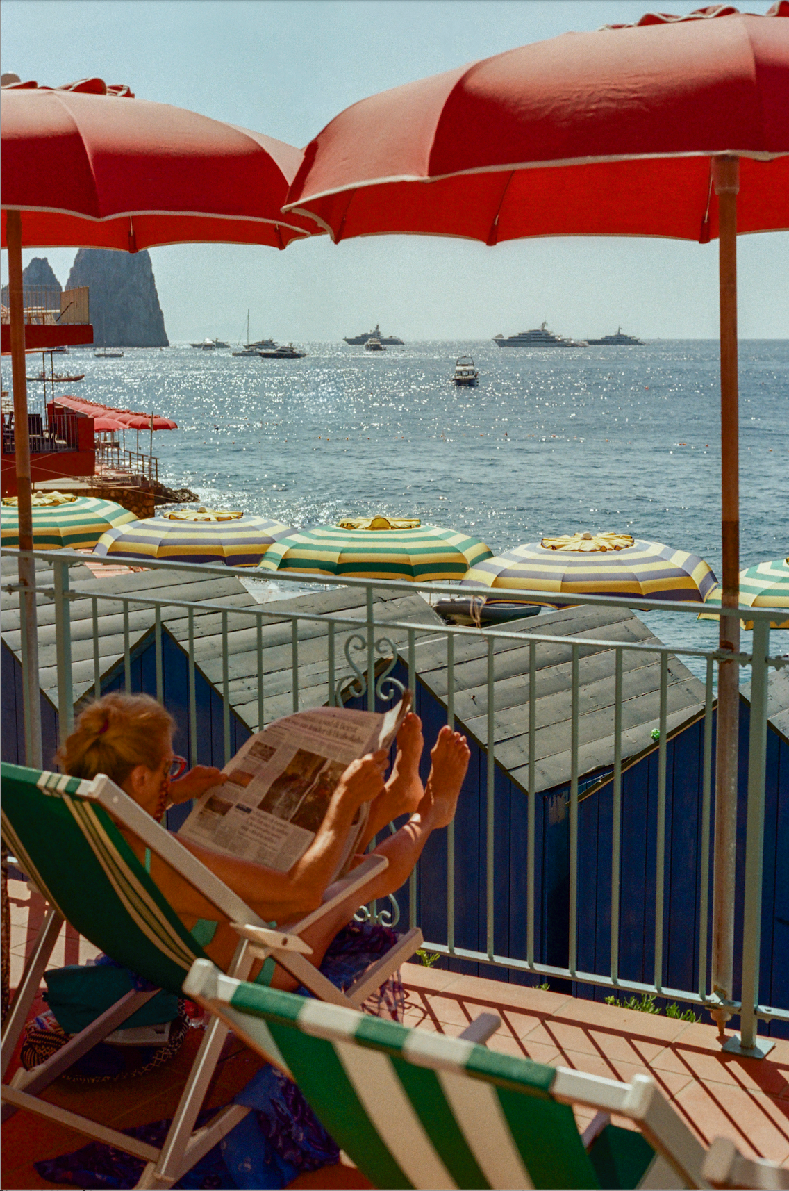 Photograph of a woman reading the newspaper on a balcony overlooking Bagni Internazionale beach club in Capri, Italy. 